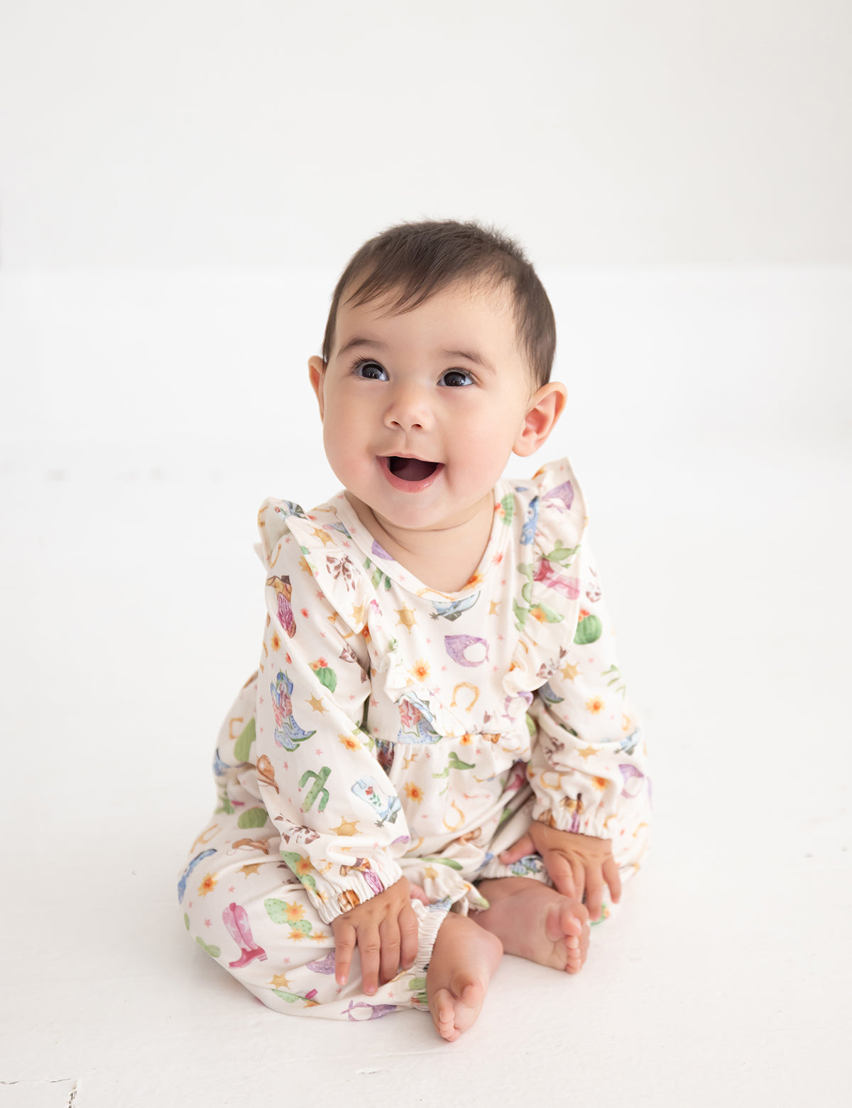 A baby sits on a white floor, smiling and looking slightly upward, dressed in the Mabel and Honey Cactus Kisses Long Sleeve Print Ruffle Neck Bubble Romper.