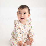 A baby sits on a white floor, smiling and looking slightly upward, dressed in the Mabel and Honey Cactus Kisses Long Sleeve Print Ruffle Neck Bubble Romper.