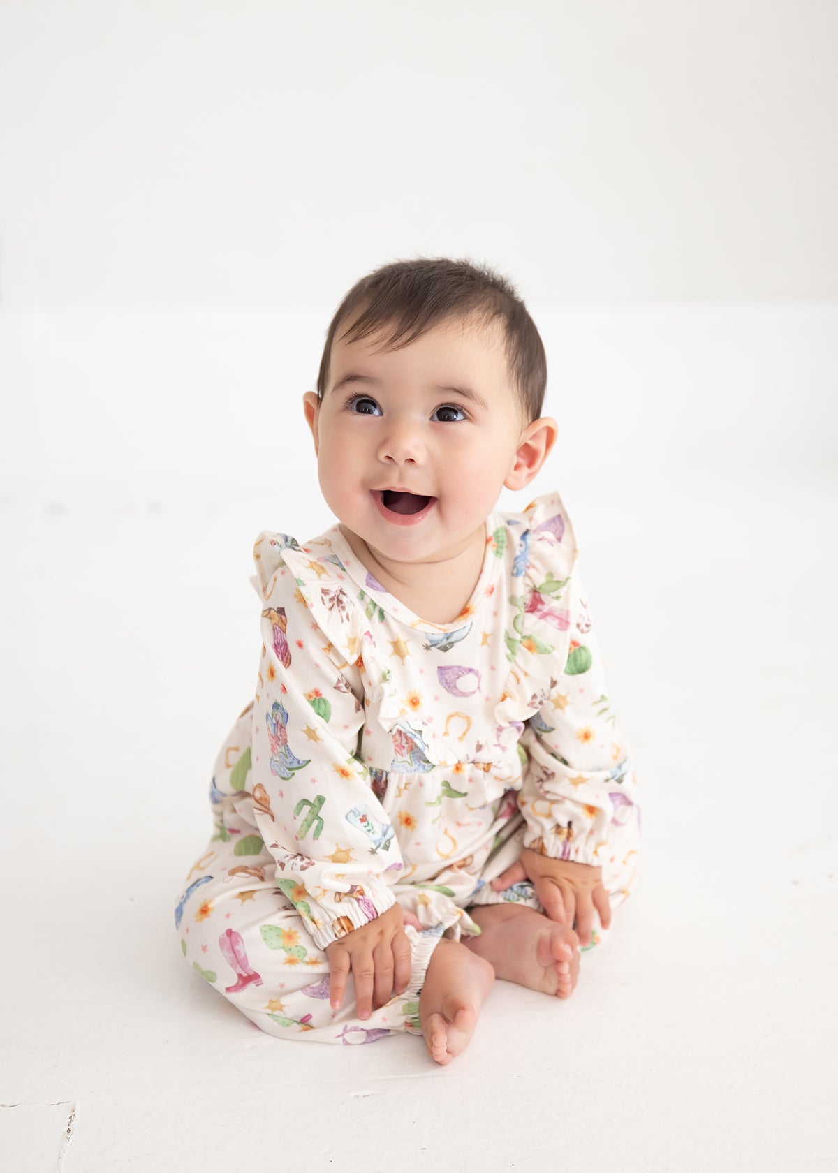 A baby sits on a white floor, smiling and looking slightly upward, dressed in the Mabel and Honey Cactus Kisses Long Sleeve Print Ruffle Neck Bubble Romper.