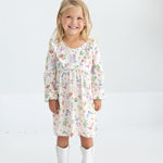 A young girl with blonde hair smiles while wearing the Mabel and Honey Cactus Kisses Long Sleeve Western Print Ruffle Yoke Dress and white cowboy boots, standing against a plain white background.