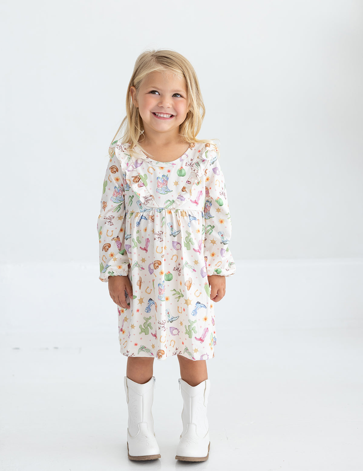 A young girl with blonde hair smiles while wearing the Mabel and Honey Cactus Kisses Long Sleeve Western Print Ruffle Yoke Dress and white cowboy boots, standing against a plain white background.