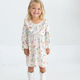 A young girl with blonde hair smiles while wearing the Mabel and Honey Cactus Kisses Long Sleeve Western Print Ruffle Yoke Dress and white cowboy boots, standing against a plain white background.