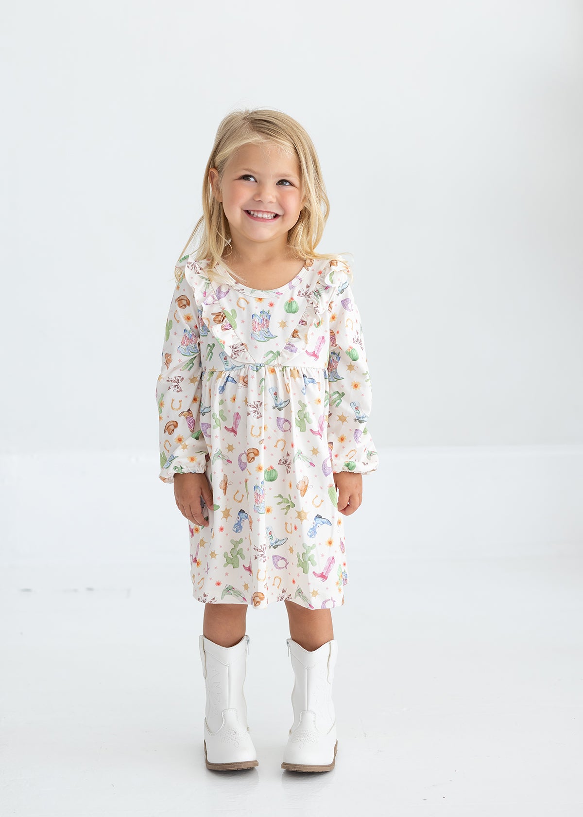 A young girl with blonde hair smiles while wearing the Mabel and Honey Cactus Kisses Long Sleeve Western Print Ruffle Yoke Dress and white cowboy boots, standing against a plain white background.