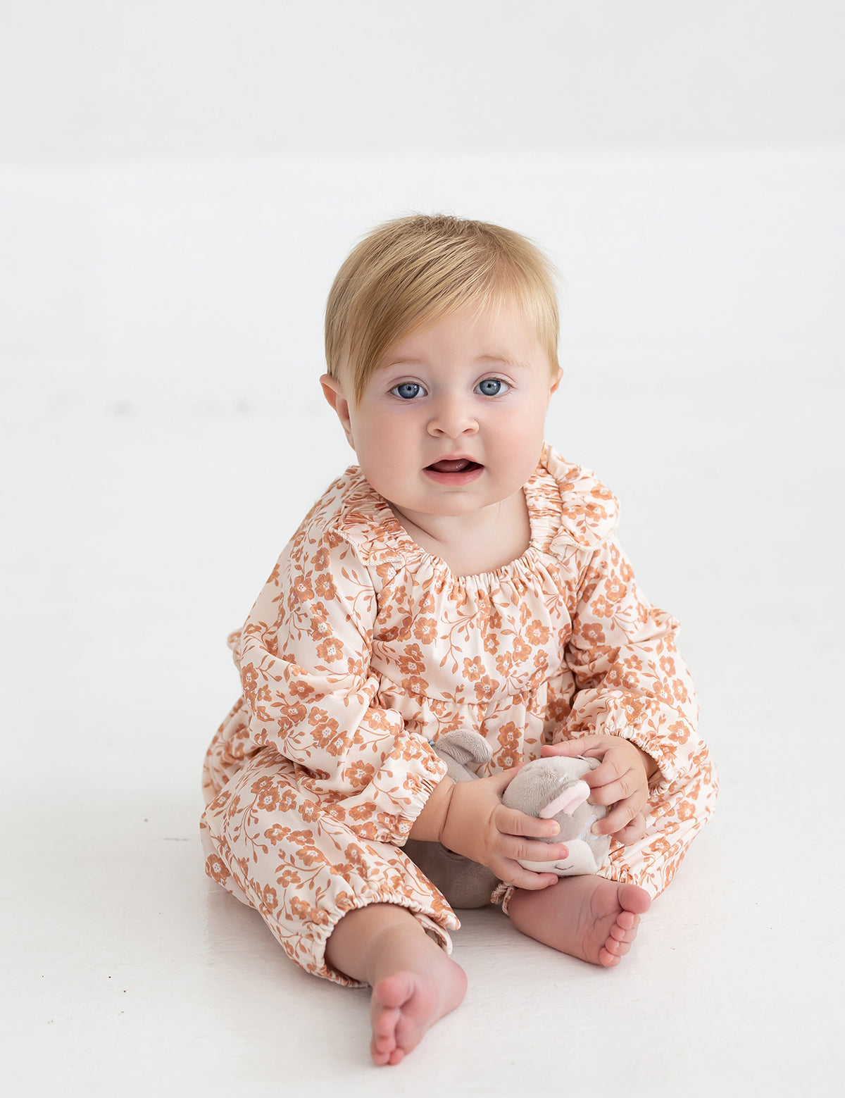 A baby with blonde hair and blue eyes sits barefoot on a white floor, wearing the Mabel and Honey Honey Petals Long Sleeve Floral Ruffle Collar Straight Leg Romper, and holding a small stuffed toy while looking toward the camera.