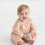 A baby with blonde hair and blue eyes sits barefoot on a white floor, wearing the Mabel and Honey Honey Petals Long Sleeve Floral Ruffle Collar Straight Leg Romper, and holding a small stuffed toy while looking toward the camera.