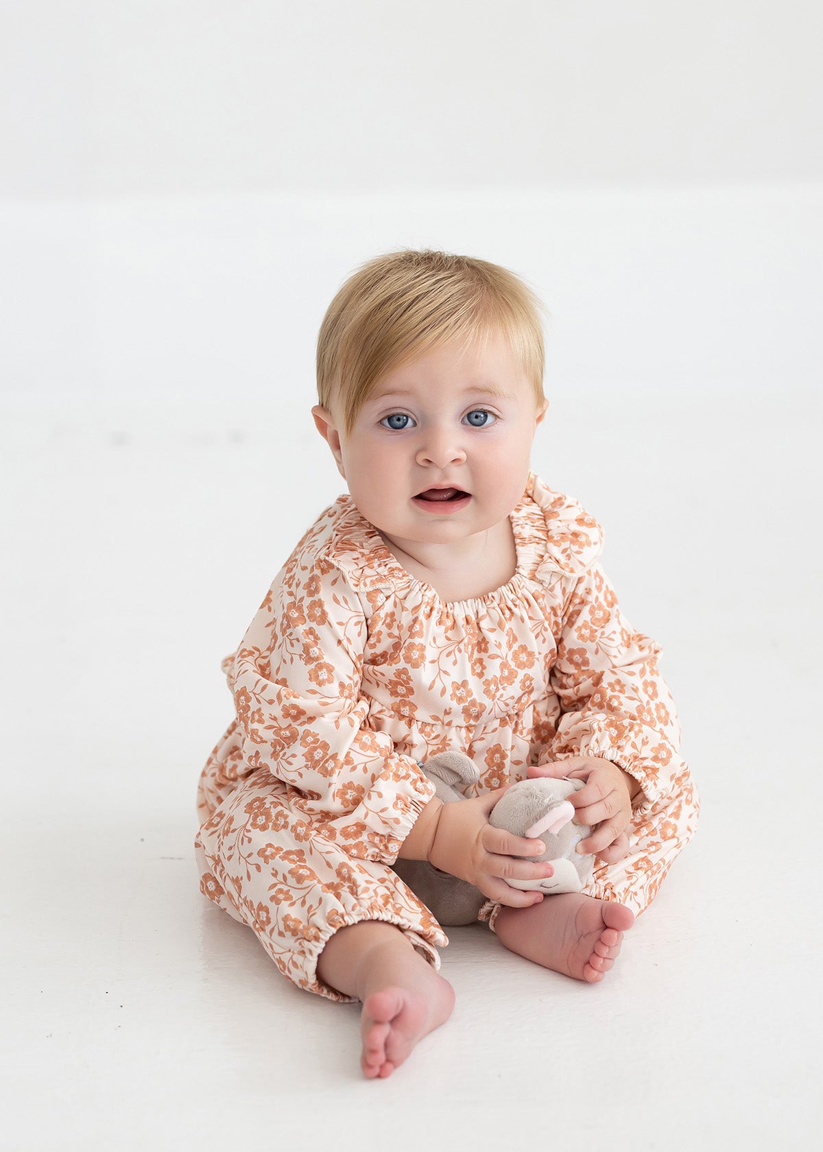 A baby with blonde hair and blue eyes sits barefoot on a white floor, wearing the Mabel and Honey Honey Petals Long Sleeve Floral Ruffle Collar Straight Leg Romper, and holding a small stuffed toy while looking toward the camera.