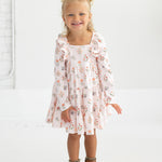 A smiling young girl with blonde hair in pigtails wears the Mabel and Honey “Amelia Rose” Long Sleeve Floral Ruffle Trim Tiered Dress, paired with brown ankle boots, standing against a plain white background.