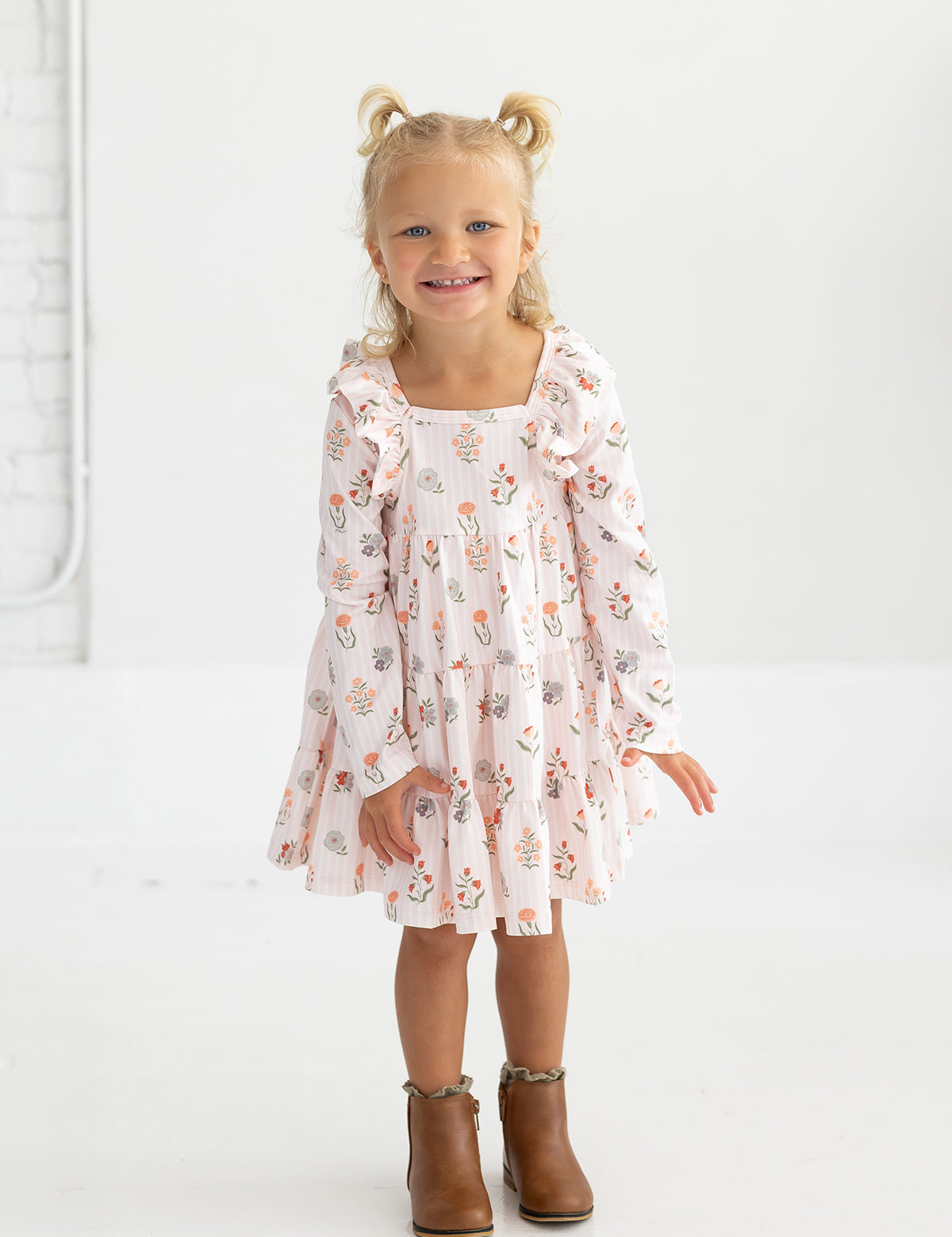 A smiling young girl with blonde hair in pigtails wears the Mabel and Honey “Amelia Rose” Long Sleeve Floral Ruffle Trim Tiered Dress, paired with brown ankle boots, standing against a plain white background.