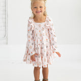 A smiling young girl with blonde hair in pigtails wears the Mabel and Honey “Amelia Rose” Long Sleeve Floral Ruffle Trim Tiered Dress, paired with brown ankle boots, standing against a plain white background.