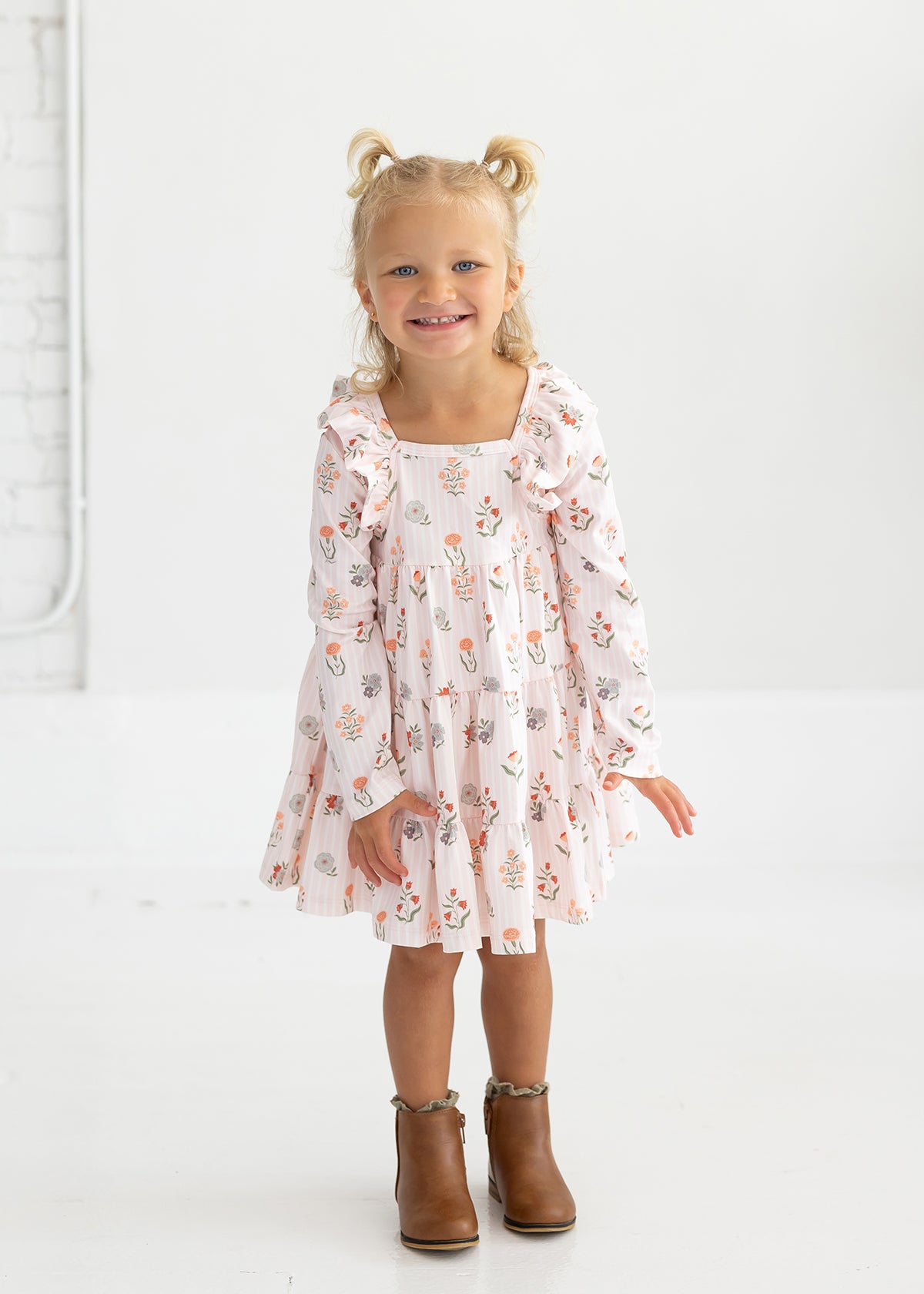 A smiling young girl with blonde hair in pigtails wears the Mabel and Honey “Amelia Rose” Long Sleeve Floral Ruffle Trim Tiered Dress, paired with brown ankle boots, standing against a plain white background.