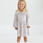 A young child with long blonde hair smiles while standing in the Mabel and Honey Soft Meadow Long Sleeve Floral Ruffle Collar Empire Waist Dress, paired with white socks and white shoes against a plain, light background.