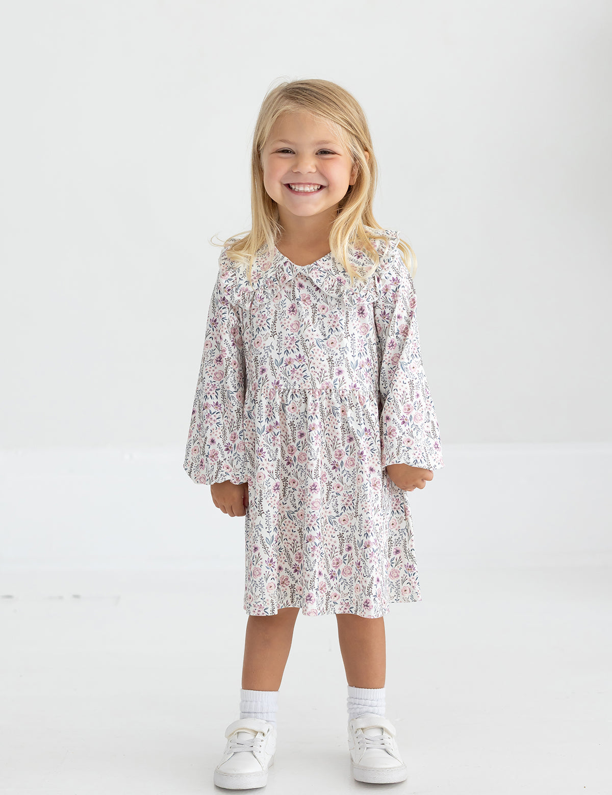 A young child with long blonde hair smiles while standing in the Mabel and Honey Soft Meadow Long Sleeve Floral Ruffle Collar Empire Waist Dress, paired with white socks and white shoes against a plain, light background.