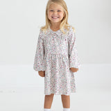 A young child with long blonde hair smiles while standing in the Mabel and Honey Soft Meadow Long Sleeve Floral Ruffle Collar Empire Waist Dress, paired with white socks and white shoes against a plain, light background.