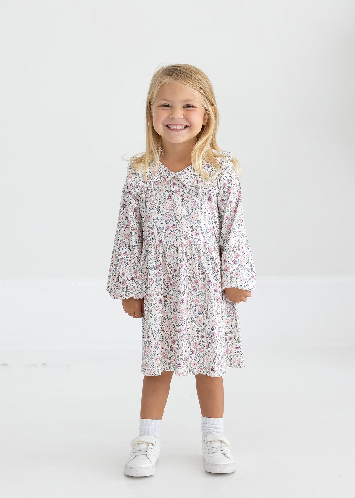 A young child with long blonde hair smiles while standing in the Mabel and Honey Soft Meadow Long Sleeve Floral Ruffle Collar Empire Waist Dress, paired with white socks and white shoes against a plain, light background.