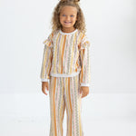 A young child with curly hair styled in two buns stands smiling, dressed in Mabel and Honey’s Buttercup Blossom Striped Elastic Waist Wide Leg Pants and a coordinated long-sleeve top, both crafted from a soft cotton blend with a colorful vertical striped floral print. She completes the look with white shoes against a plain white background.
