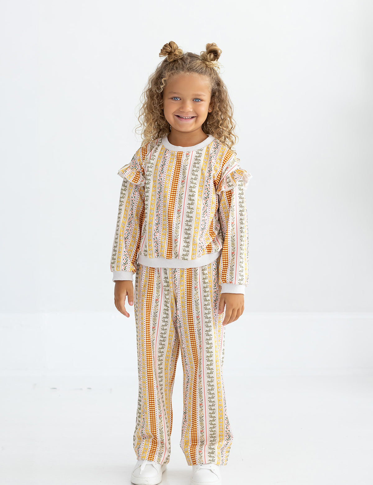 A young child with curly hair styled in two buns stands smiling, dressed in Mabel and Honey’s Buttercup Blossom Striped Elastic Waist Wide Leg Pants and a coordinated long-sleeve top, both crafted from a soft cotton blend with a colorful vertical striped floral print. She completes the look with white shoes against a plain white background.