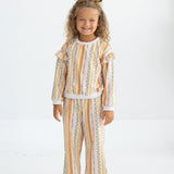 A young child with curly hair styled in two buns stands smiling, dressed in Mabel and Honey’s Buttercup Blossom Striped Elastic Waist Wide Leg Pants and a coordinated long-sleeve top, both crafted from a soft cotton blend with a colorful vertical striped floral print. She completes the look with white shoes against a plain white background.