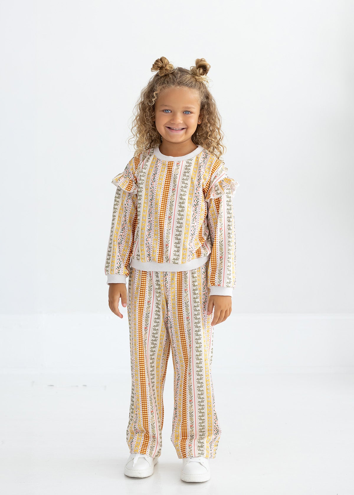 A young child with curly hair styled in two buns stands smiling, dressed in Mabel and Honey’s Buttercup Blossom Striped Elastic Waist Wide Leg Pants and a coordinated long-sleeve top, both crafted from a soft cotton blend with a colorful vertical striped floral print. She completes the look with white shoes against a plain white background.