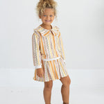 A young child with curly blonde hair smiles while standing against a plain white background, wearing the Mabel and Honey Buttercup Blossom Long Sleeve Striped Floral Ruffle Collar Sweater paired with brown ankle boots.