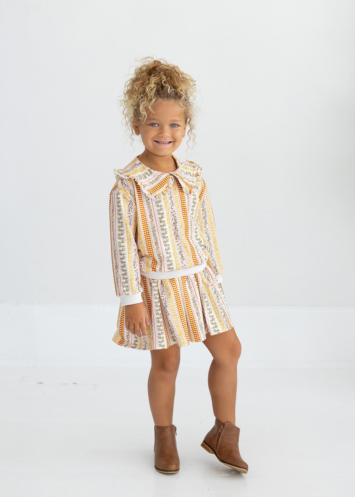 A young child with curly blonde hair smiles while standing against a plain white background, wearing the Mabel and Honey Buttercup Blossom Long Sleeve Striped Floral Ruffle Collar Sweater paired with brown ankle boots.