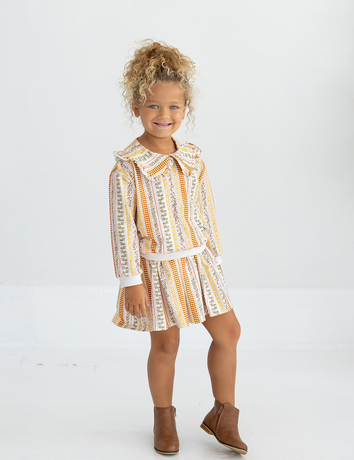 A young child with curly blonde hair smiles while wearing the Buttercup Blossom Striped Floral Pleated Elastic Waist Skirt from Mabel and Honey, paired with a collared, patterned dress and brown ankle boots, standing against a plain white background.