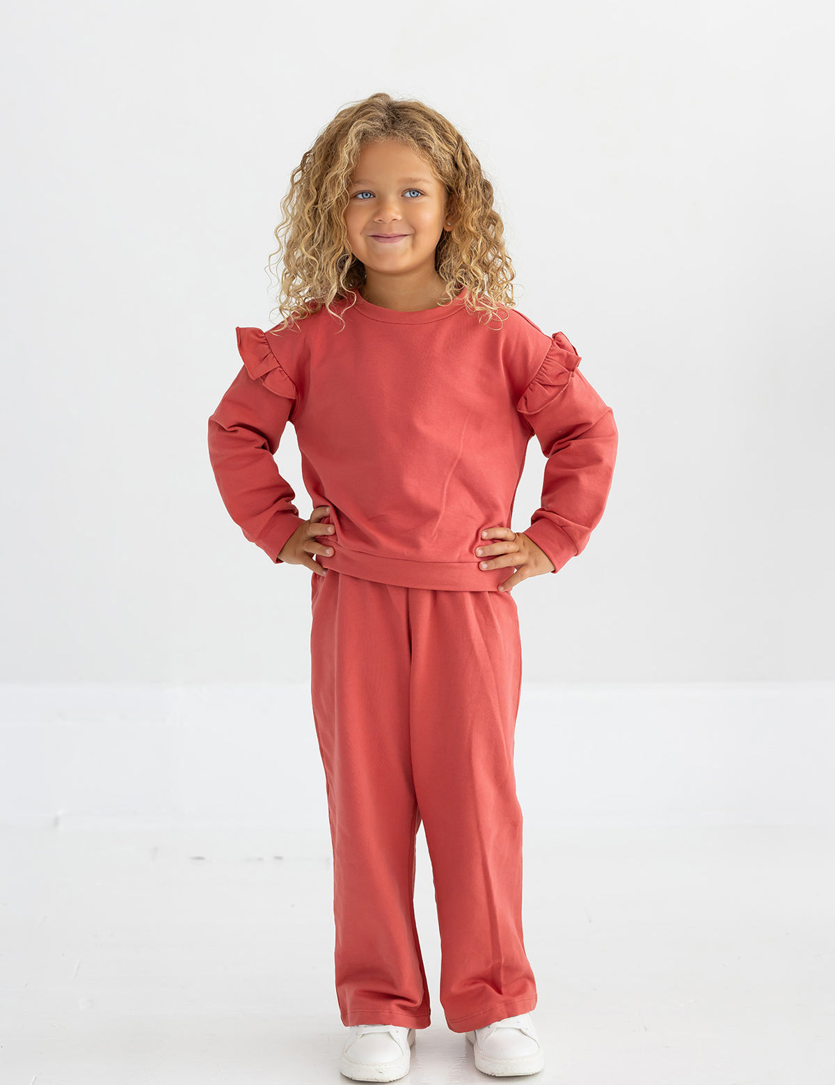 A young child with curly blonde hair wears the Mabel and Honey Red Rosie Solid Wide Leg Elastic Waist Sweatpants, paired with a matching long-sleeve ruffled top and white shoes, standing and smiling against a plain white background.