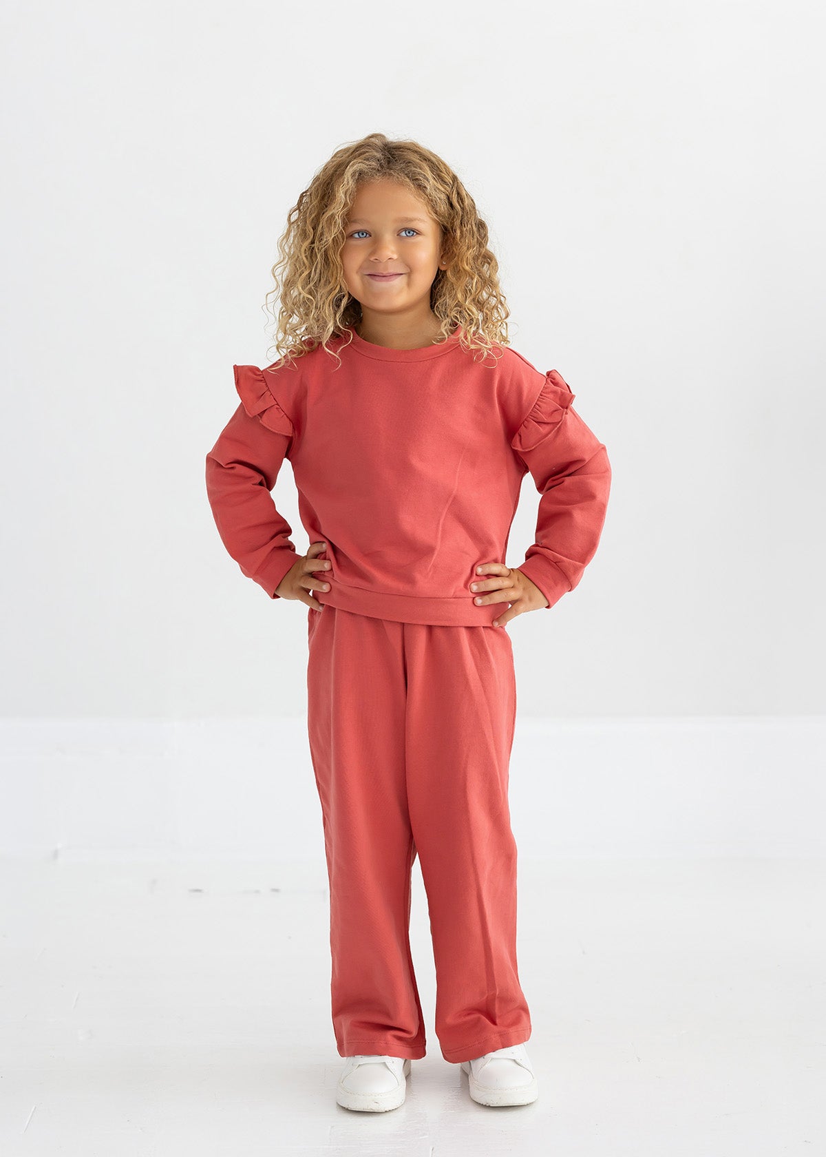 A young child with curly blonde hair wears the Mabel and Honey Red Rosie Solid Wide Leg Elastic Waist Sweatpants, paired with a matching long-sleeve ruffled top and white shoes, standing and smiling against a plain white background.