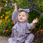 A baby in the Char Long Sleeve Plaid Ruffle Collar Bubble Romper by Mabel and Honey—crafted from soft brushed cotton flannel in a lavender plaid, featuring a ruffled collar and matching headband—sits barefoot on a sidewalk, smiling with both hands raised. Colorful flowers and greenery fill the background.
