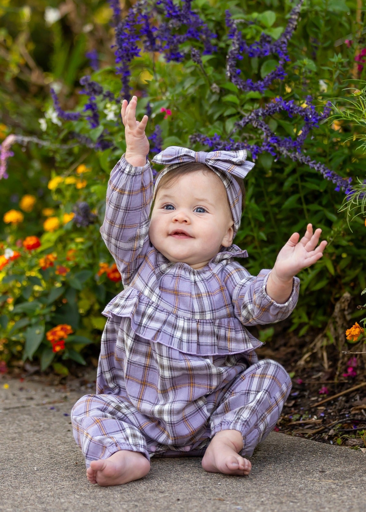 A baby in the Char Long Sleeve Plaid Ruffle Collar Bubble Romper by Mabel and Honey—crafted from soft brushed cotton flannel in a lavender plaid, featuring a ruffled collar and matching headband—sits barefoot on a sidewalk, smiling with both hands raised. Colorful flowers and greenery fill the background.