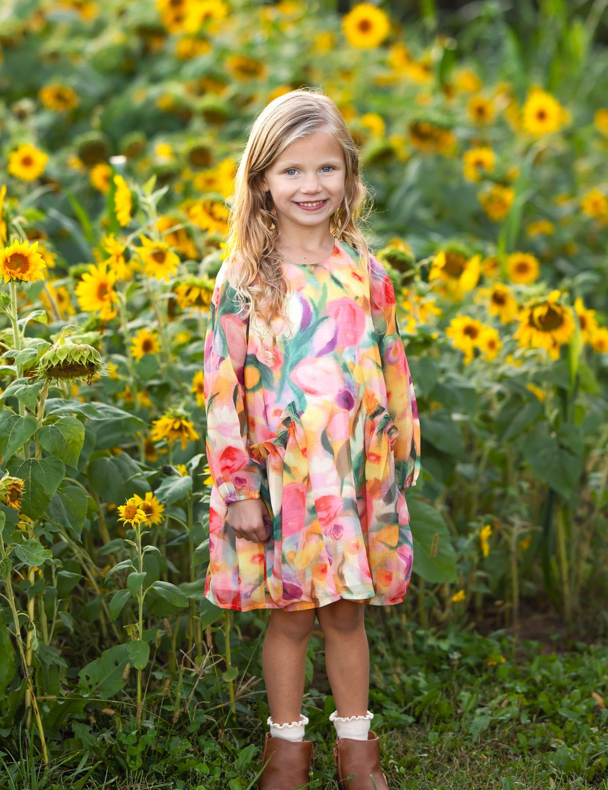 A young girl with long blonde hair smiles as she stands in front of a field of blooming sunflowers. She wears the Mabel and Honey Watercolor Whimsey Long Sleeve Floral Tiered Ruffle Waist Dress, featuring vibrant watercolor floral patterns, paired with brown boots.
