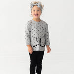 A smiling toddler stands against a white background, wearing the Mabel and Honey Petal Pockets Long Sleeve Floral Scalloped Pocket Top and Legging Set, paired with white shoes and a matching gray bow headband.