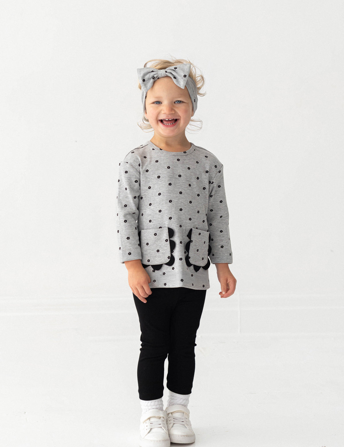 A smiling toddler stands against a white background, wearing the Mabel and Honey Petal Pockets Long Sleeve Floral Scalloped Pocket Top and Legging Set, paired with white shoes and a matching gray bow headband.