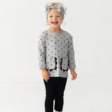 A smiling toddler stands against a white background, wearing the Mabel and Honey Petal Pockets Long Sleeve Floral Scalloped Pocket Top and Legging Set, paired with white shoes and a matching gray bow headband.