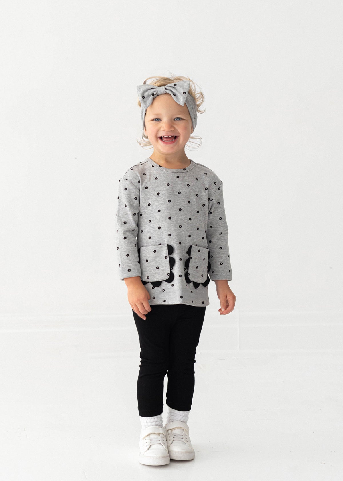 A smiling toddler stands against a white background, wearing the Mabel and Honey Petal Pockets Long Sleeve Floral Scalloped Pocket Top and Legging Set, paired with white shoes and a matching gray bow headband.