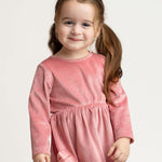 A young girl with long brown hair in pigtails stands and smiles at the camera against a plain light background, wearing the Mabel and Honey Bowtiful Blush Long Sleeve Solid Ribbed Bow Trim A Line Dress.