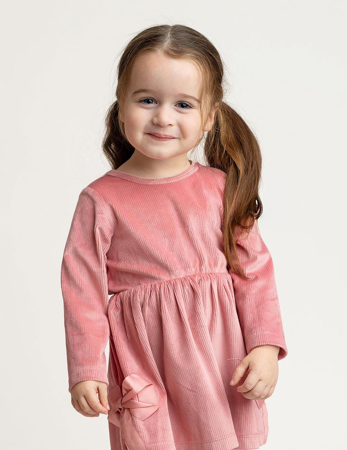 A young girl with long brown hair in pigtails stands and smiles at the camera against a plain light background, wearing the Mabel and Honey Bowtiful Blush Long Sleeve Solid Ribbed Bow Trim A Line Dress.