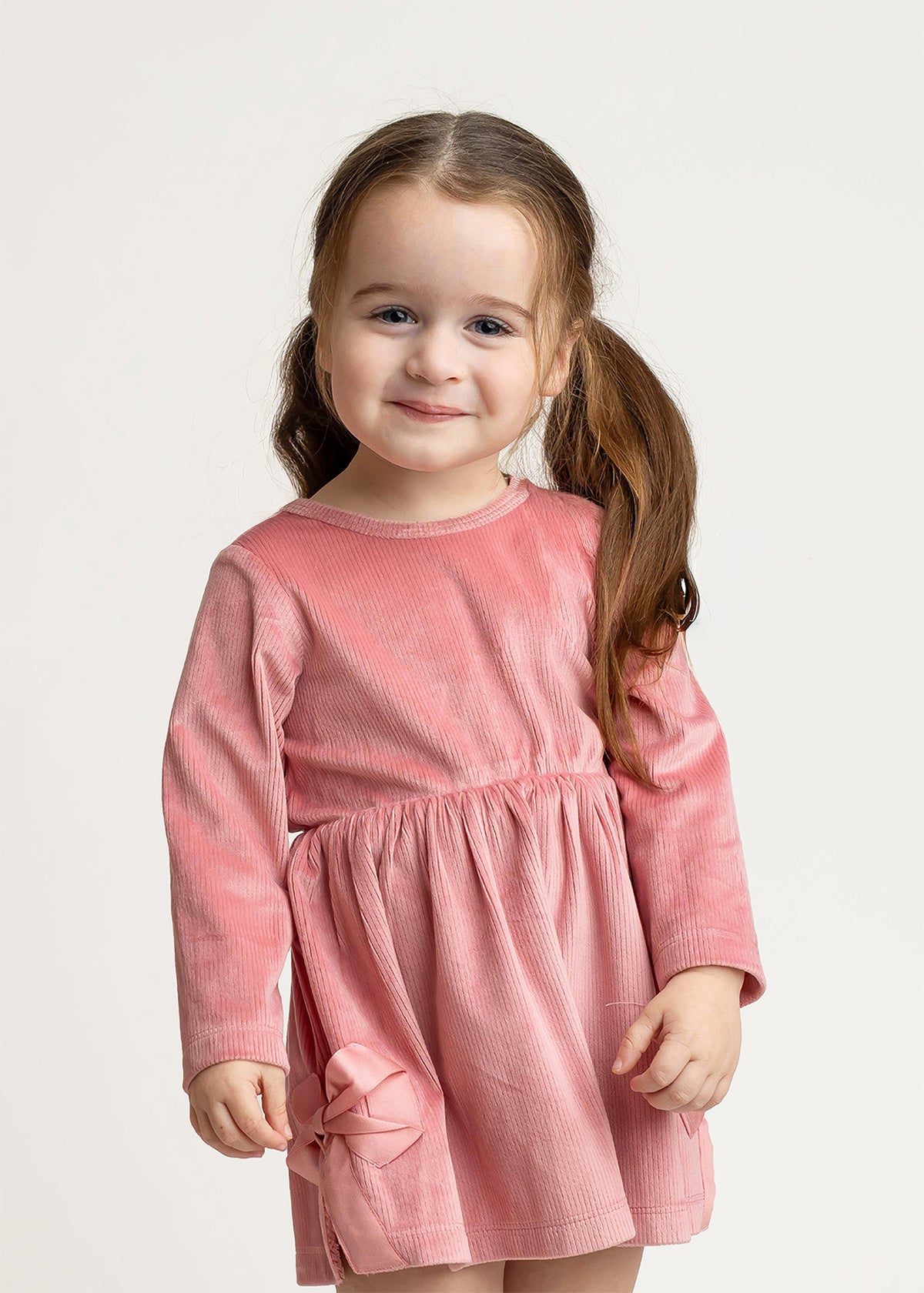 A young girl with long brown hair in pigtails stands and smiles at the camera against a plain light background, wearing the Mabel and Honey Bowtiful Blush Long Sleeve Solid Ribbed Bow Trim A Line Dress.