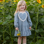 A young girl with blonde hair stands smiling in a sunflower field, holding a sunflower and wearing light-colored boots. She is dressed in the Mabel and Honey Dolly Long Sleeve Floral Lace Heart Applique Tiered Dress.