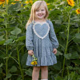 A young girl with blonde hair stands smiling in a sunflower field, holding a sunflower and wearing light-colored boots. She is dressed in the Mabel and Honey Dolly Long Sleeve Floral Lace Heart Applique Tiered Dress.