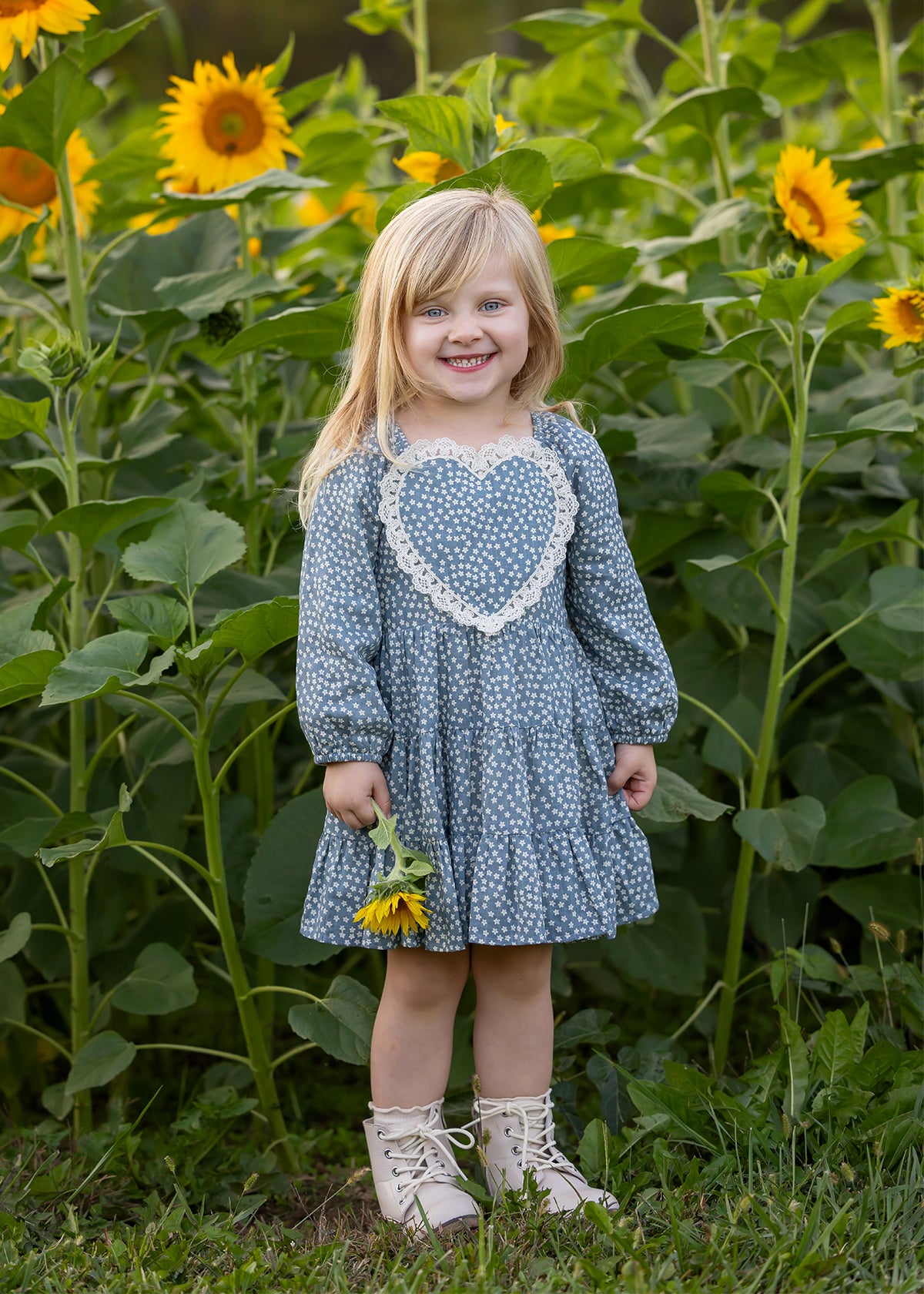 A young girl with blonde hair stands smiling in a sunflower field, holding a sunflower and wearing light-colored boots. She is dressed in the Mabel and Honey Dolly Long Sleeve Floral Lace Heart Applique Tiered Dress.