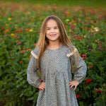 A young girl with long hair stands smiling in a flower field, her hands on her hips and colorful blossoms in the background, wearing the Mabel and Honey Dolly Long Sleeve Floral Lace Trim Tie Waist Dress.