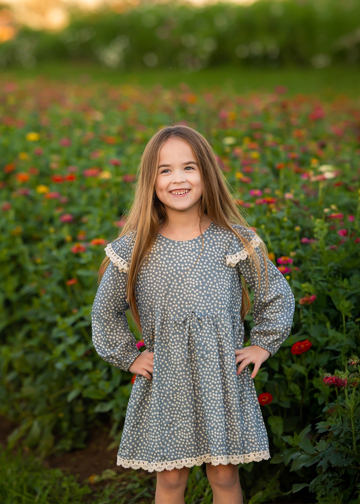 A young girl with long hair stands smiling in a flower field, her hands on her hips and colorful blossoms in the background, wearing the Mabel and Honey Dolly Long Sleeve Floral Lace Trim Tie Waist Dress.