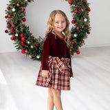 A young girl wears the Isobella and Chloe Holiday Plaid Long Sleeve Velvet Bodice Bow Tie Waist Fit and Flare Dress, smiling as she stands in front of a large Christmas wreath adorned with red ornaments in a bright room.