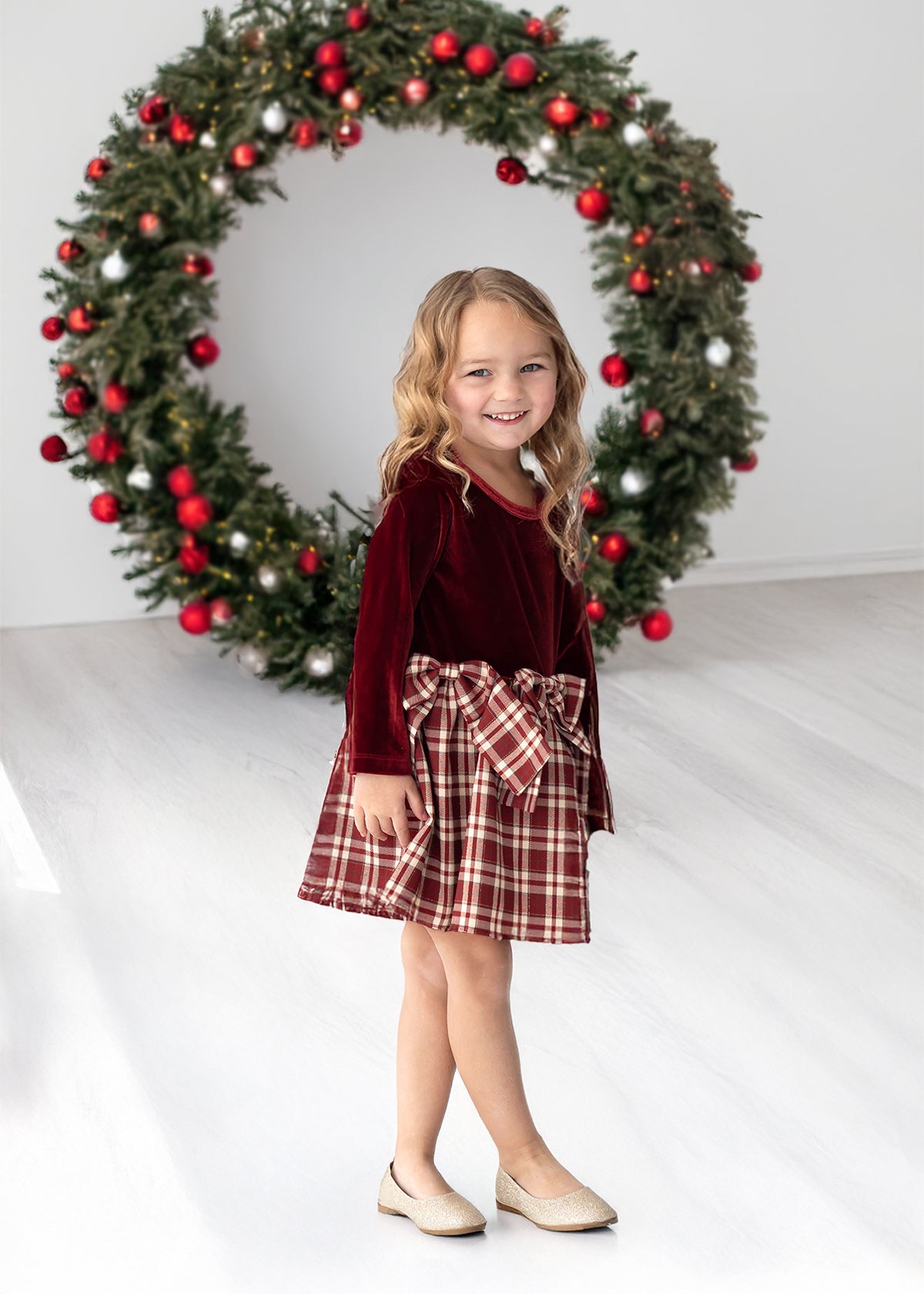 A young girl wears the Isobella and Chloe Holiday Plaid Long Sleeve Velvet Bodice Bow Tie Waist Fit and Flare Dress, smiling as she stands in front of a large Christmas wreath adorned with red ornaments in a bright room.