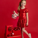 A young girl wearing the Isobella and Chloe Razzle Dazzle Short Sleeve Metallic Tiered Drop Waist Dress stands smiling with one leg crossed, holding a large striped ornament. She is surrounded by red-wrapped gifts and ornaments against a red background.