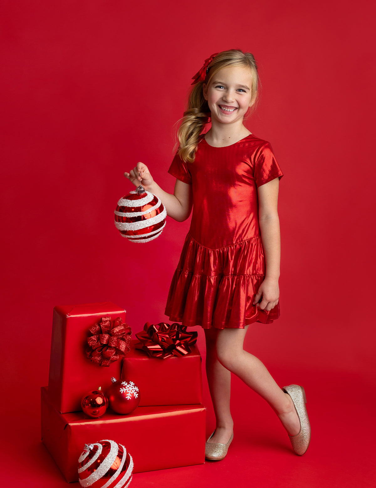 A young girl wearing the Isobella and Chloe Razzle Dazzle Short Sleeve Metallic Tiered Drop Waist Dress stands smiling with one leg crossed, holding a large striped ornament. She is surrounded by red-wrapped gifts and ornaments against a red background.