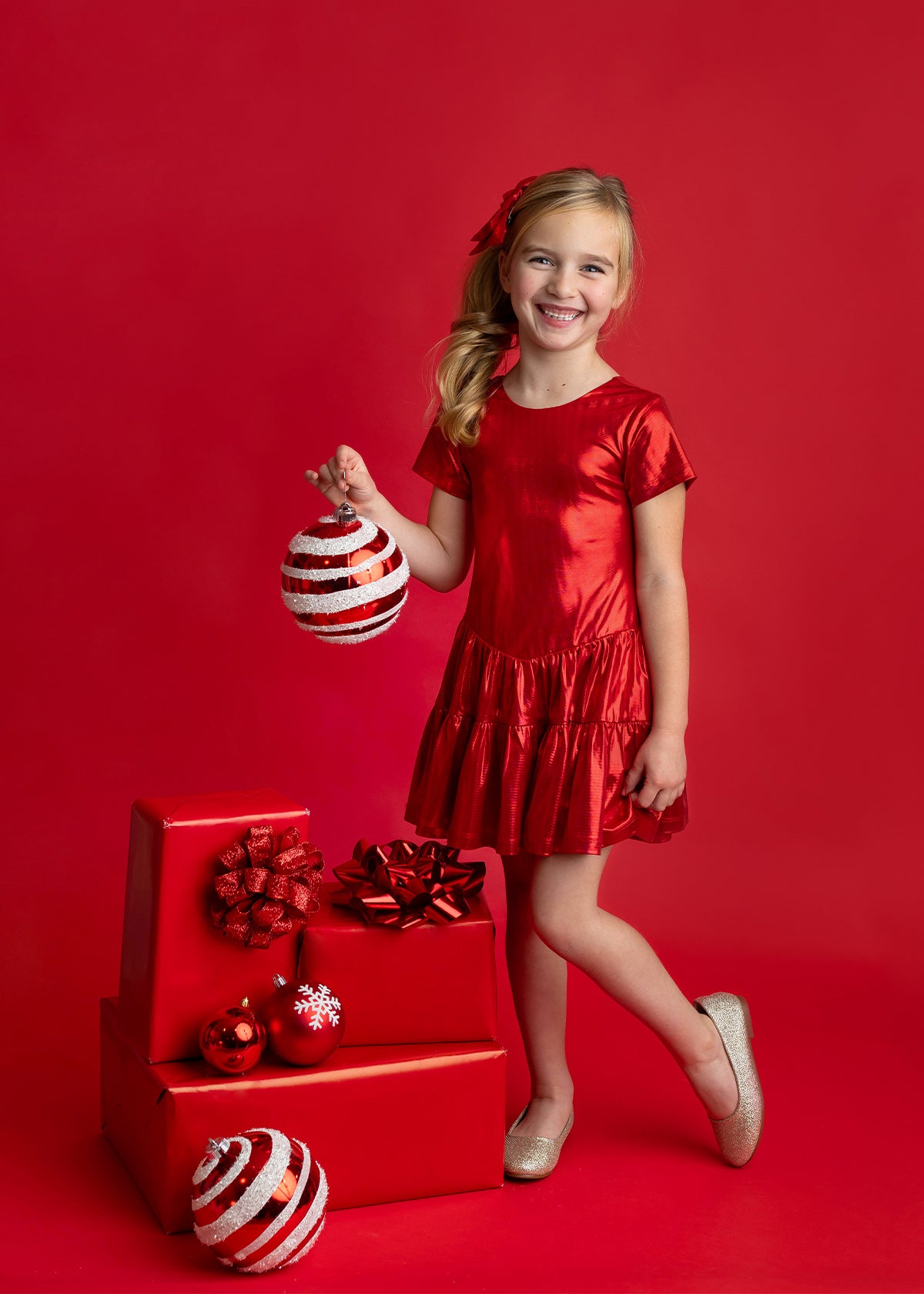 A young girl wearing the Isobella and Chloe Razzle Dazzle Short Sleeve Metallic Tiered Drop Waist Dress stands smiling with one leg crossed, holding a large striped ornament. She is surrounded by red-wrapped gifts and ornaments against a red background.