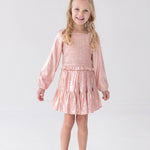 A young girl with long blonde hair wears the Rosy Glow Long Sleeve Smocked Metallic Tiered Skirt Set by Isobella and Chloe, featuring a shiny light pink crop top and matching skirt, paired with coordinating pink shoes, standing and smiling against a plain white background—an adorable special occasion outfit.