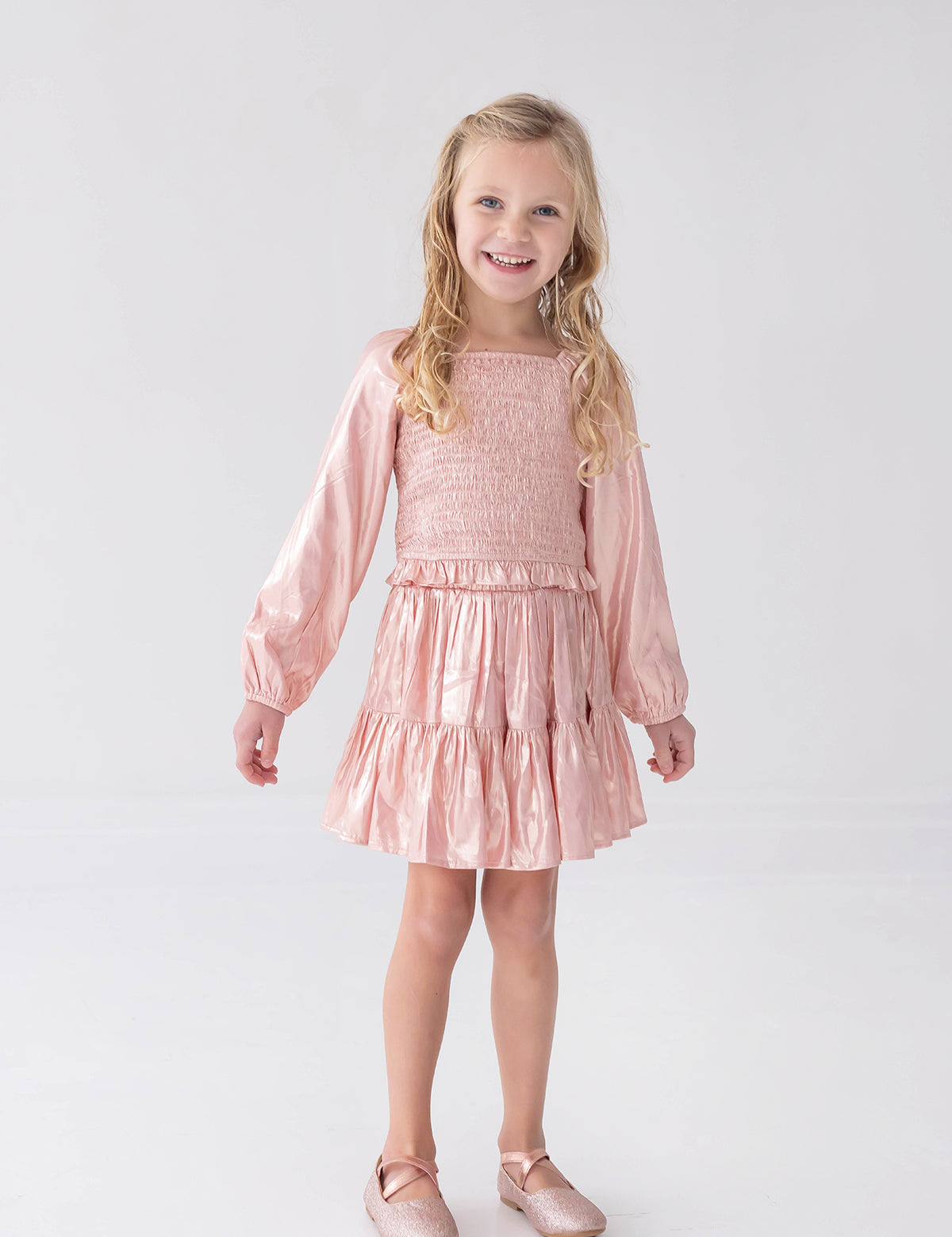 A young girl with long blonde hair wears the Rosy Glow Long Sleeve Smocked Metallic Tiered Skirt Set by Isobella and Chloe, featuring a shiny light pink crop top and matching skirt, paired with coordinating pink shoes, standing and smiling against a plain white background—an adorable special occasion outfit.