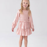 A young girl with long blonde hair wears the Rosy Glow Long Sleeve Smocked Metallic Tiered Skirt Set by Isobella and Chloe, featuring a shiny light pink crop top and matching skirt, paired with coordinating pink shoes, standing and smiling against a plain white background—an adorable special occasion outfit.